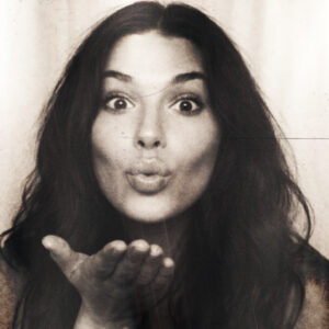 Photo of a woman blowing a kiss inside an enclosed photo booth. A vintage, analog filter is applied.